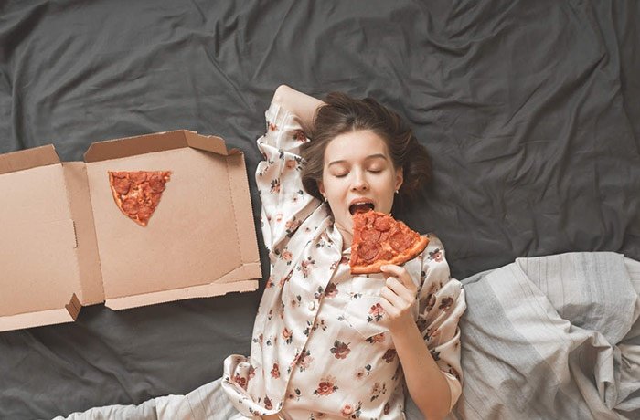 Yatağa sırtüstü uzanmış pizza yiyen bir kadın